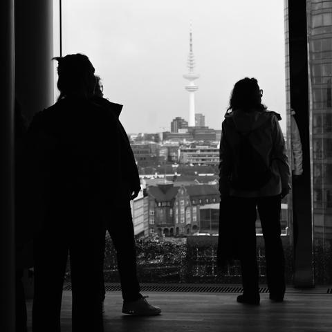 Blick aus einem Fenster. Links und Rechts zwei Menschen als schwarze Silhouetten, in der Mitte ein Fernsehturm.
