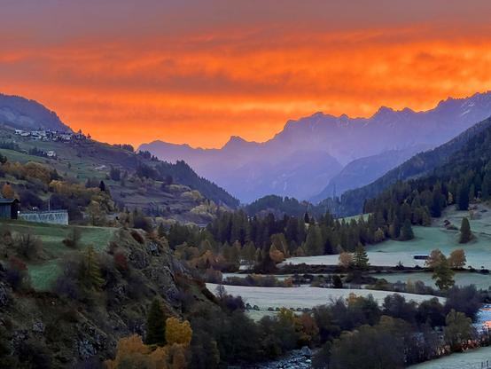 Ein spektakulärer Sonnenaufgang in den Alpen. Der Himmel leuchtet in den intensivsten Orange- und Rottönen, während die fernen Berggipfel in ein sanftes Lila getaucht sind. Im Tal liegt noch der Hauch von Frost auf den Wiesen.