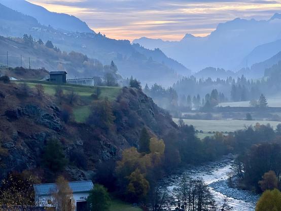 Es ist eine Landschaftsaufnahme im frühen Morgenlicht. Unten rechts fließt ein heller, steiniger Fluss durch das Bild, umgeben von Bäumen und Büschen, deren Laub teilweise schon herbstlich gelb und rotbraun gefärbt ist. Im ganz unteren Bereich links ist ein Teil eines kleinen Gebäudes mit einem Blechdach sichtbar. Der Fluss fließt durch ein breites Tal mit flachen Wiesen. Eine steile, felsige Böschung trennt den unteren Bereich von einem höheren Plateau. Das Tal ist teilweise in nebligen Dunst …
