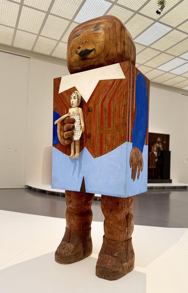 Baby Boy, 1962/63

Das Bild zeigt eine großformatige, aus Holz gefertigte Skulptur in einem Museumsraum. Die Figur wirkt wie eine stilisierte, blockartige Marionette oder Spielzeugfigur. Ihr Oberkörper ist quadratisch und bemalt, mit einer gestreiften Weste und hellblauen Akzenten. Der Kopf ist oval und trägt ein freundlich-groteskes Gesicht. An der rechten Hand hält die Figur eine kleine, stoffartige Puppe. Die Beine sind grob geschnitzt, unbemalt und enden in massiven Stiefeln. Im Hintergrund…