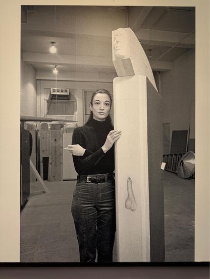 Marisol in ihrem Atelier mit der Skulptur Guy (1967/68), 1983, fotografiert von Jack Mitchell

Das schwarz-weiße Foto zeigt eine Frau in einem Atelier, die neben einer hohen, schlanken Skulptur steht und ihre Hand auf diese legt. Sie trägt einen dunklen Rollkragenpullover und eine dunkle Hose, ihre Haltung ist ruhig und bestimmt. Die Skulptur ist oben mit einer geschnitzten, gesichtslosen Maskenform versehen und weist auf der Vorderseite eine stilisierte Abbildung von männlichen Genitalien auf.…