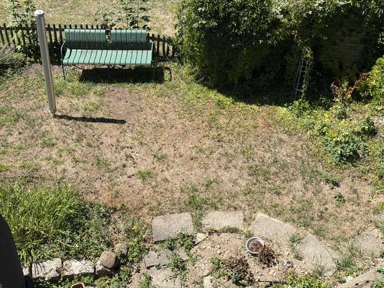 Das Bild zeigt die Vogelperspektive eines sonnigen Gartens mit überwiegend vertrocknetem, braunem Rasen. Im oberen linken Bereich steht eine grüne Sitzbank vor einem niedrigen Holzzaun, daneben ragt ein silberner Metallpfosten empor. Auf der rechten Seite wachsen dichte, grüne Büsche und Kletterpflanzen. Im Vordergrund befinden sich einige graue Steinplatten, Pflastersteine sowie ein kleiner Blumentopf auf der erdigen Fläche. Das Motiv wirkt stark vom Sommer geprägt.
